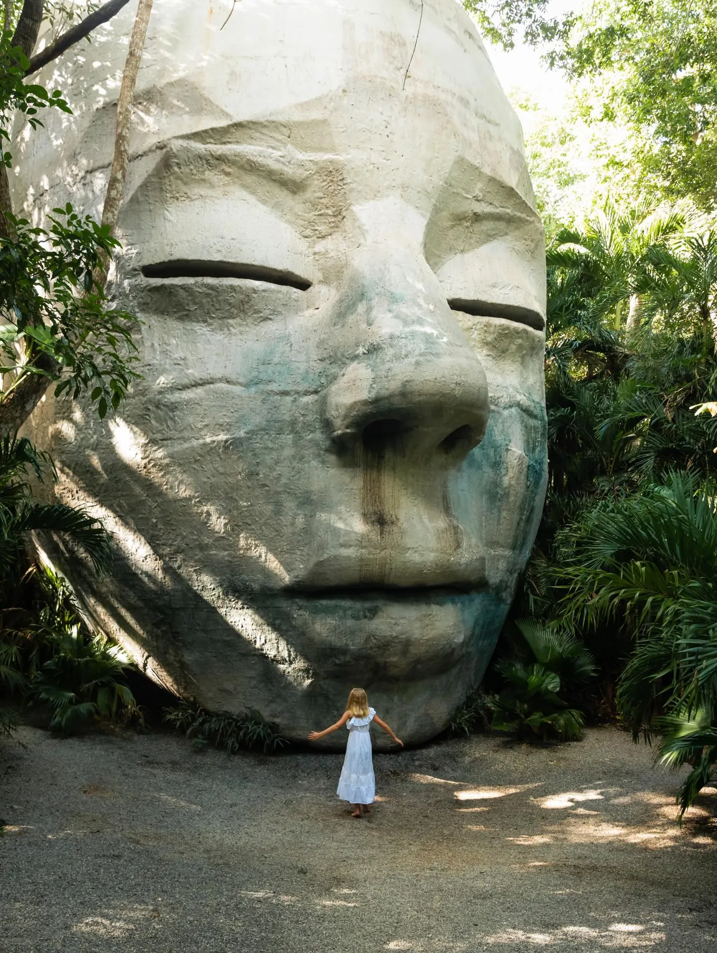 Una cabeza humanoide de piedra de 14 metros con los ojos cerrados, en la selva, con una visitante de blanco a sus pies